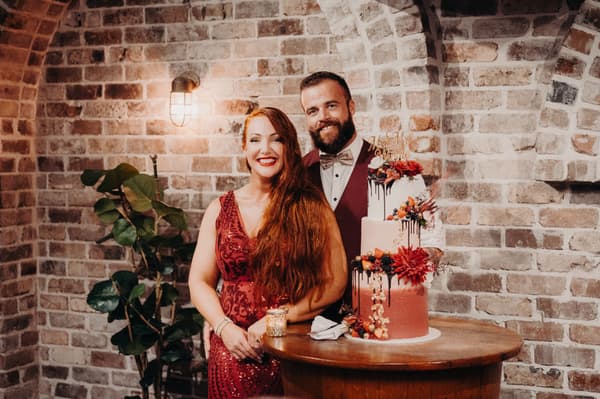 The bride in a red dress and the groom in a burgundy vest and bow tie stand behind a three-tier wedding cake decorated with flowers and berries at Sandstone Point Hotel — The Cellar.