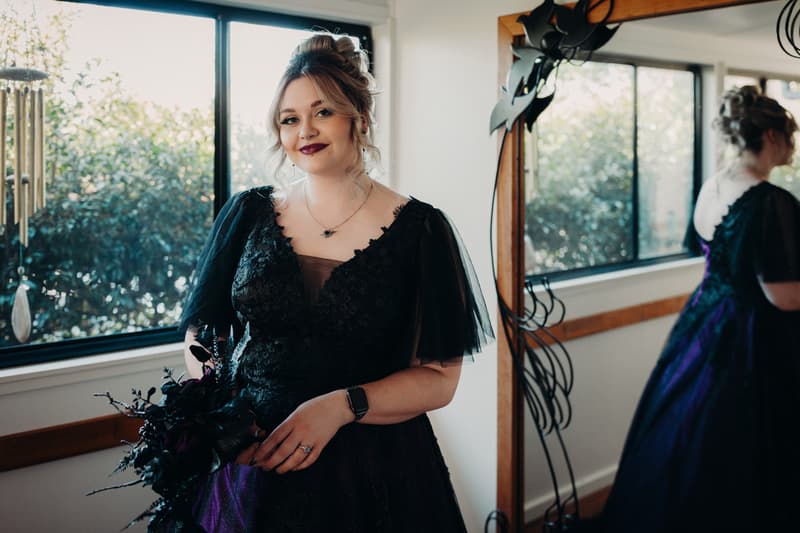 The bride Renae stands indoors near a window at Ocean View Estates, holding a dark bouquet and wearing a black lace dress with sheer sleeves, her reflection visible in a nearby mirror.
