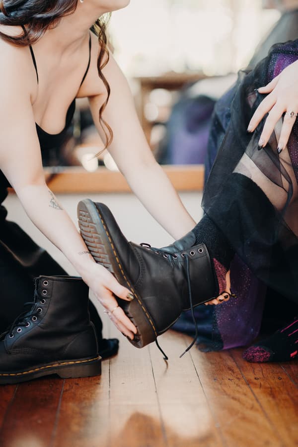 The bride helps the groom put on black lace-up boots indoors on a wooden floor.