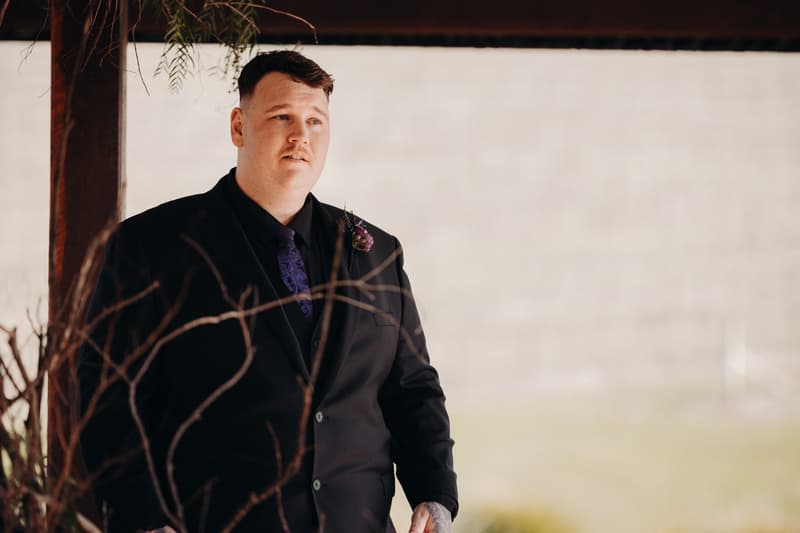 The groom Adam stands under a wooden structure at Ocean View Estates — On The Lake, wearing a black suit with a purple tie and boutonniere.