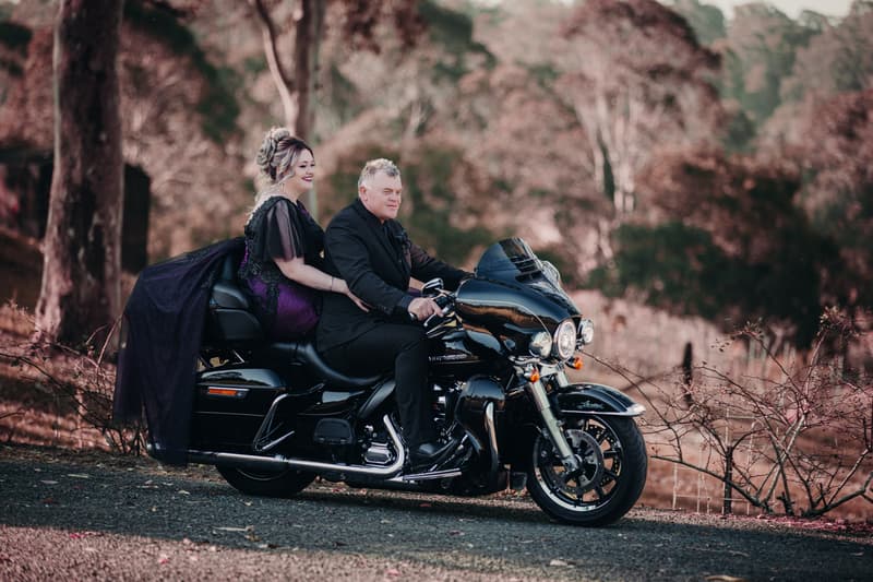 The bride and groom sit together on a black motorcycle outdoors at Ocean View Estates — On The Lake, surrounded by trees and natural scenery.