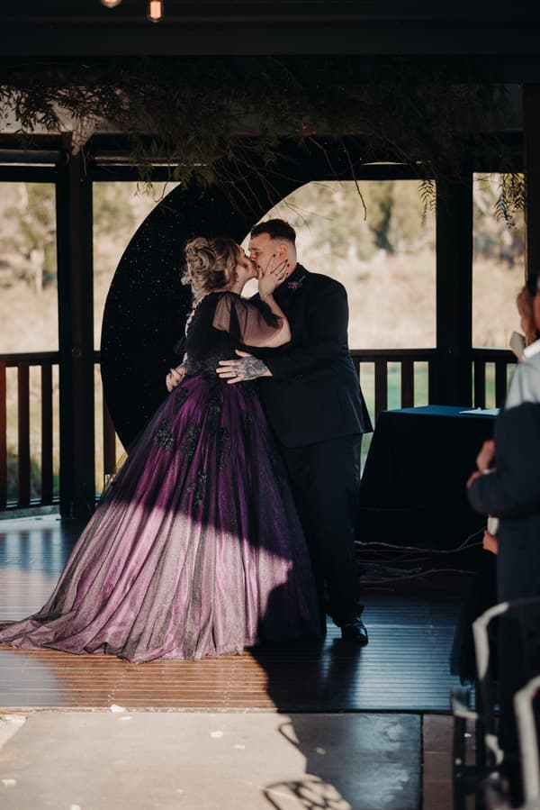 Renae and Adam share a kiss on the ceremony stage at Ocean View Estates — On The Lake, with Renae wearing a purple and black gown and Adam in a black suit.