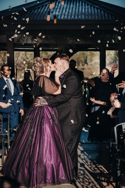 The bride and groom share a kiss at the ceremony stage at Ocean View Estates — On The Lake, surrounded by guests throwing flower petals.