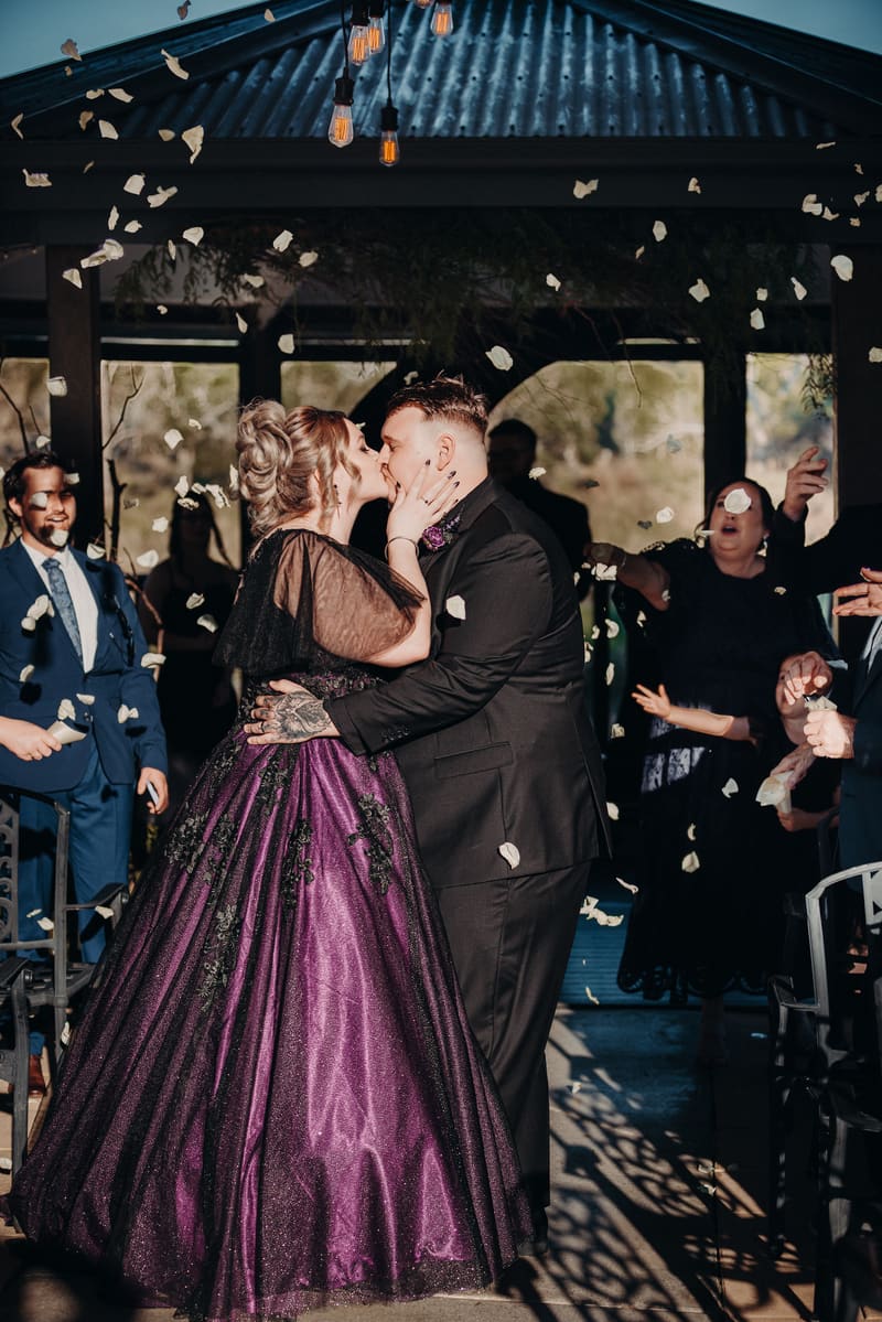 The bride and groom share a kiss at the ceremony stage at Ocean View Estates — On The Lake, surrounded by guests throwing flower petals.