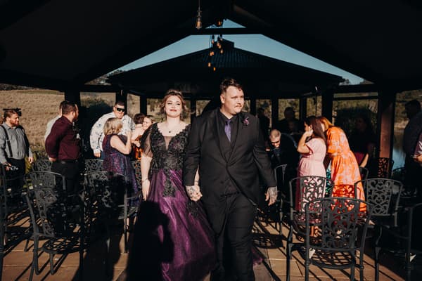 Bride Renae in a purple and black gown and groom Adam in a black suit walk hand in hand down the aisle at Ocean View Estates — On The Lake, with guests seated and standing around them under a pavilion.
