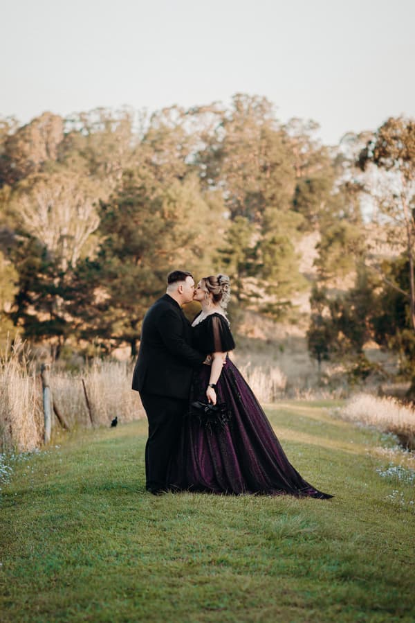 Renae and Adam share a kiss during their couple portraits session at Ocean View Estates, standing on a grassy path with trees in the background. Renae wears a dark purple gown and holds a bouquet, while Adam is dressed in a black suit.
