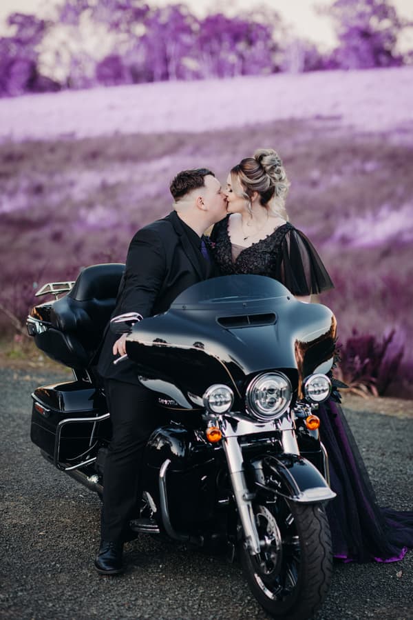 Renae and Adam kiss while seated on a black motorcycle at Ocean View Estates with a purple-hued natural background.