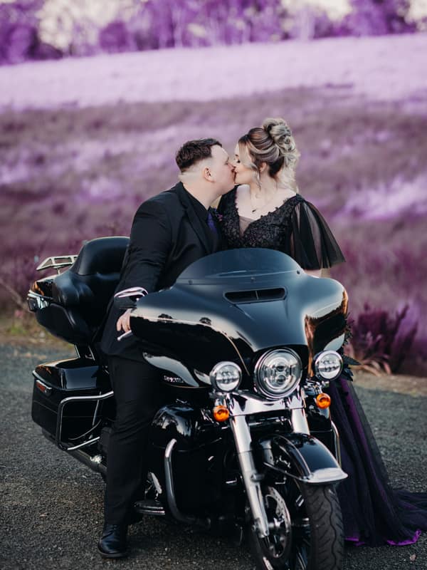 Renae and Adam kiss while seated on a black motorcycle at Ocean View Estates with a purple-hued natural background.