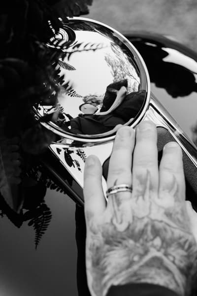 Reflection of bride Renae and groom Adam kissing is visible in a shiny car side mirror, with a tattooed hand wearing a wedding ring resting on the car near the mirror at Ocean View Estates.