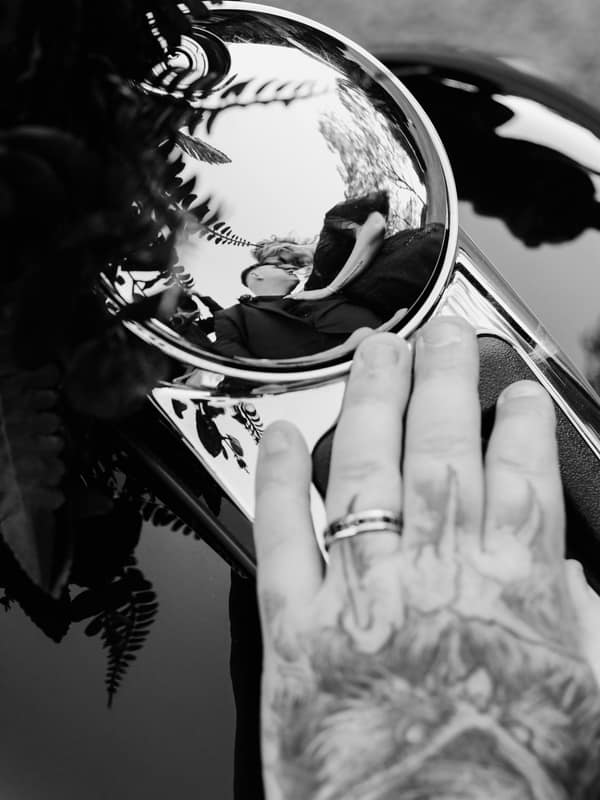 Reflection of bride Renae and groom Adam kissing is visible in a shiny car side mirror, with a tattooed hand wearing a wedding ring resting on the car near the mirror at Ocean View Estates.