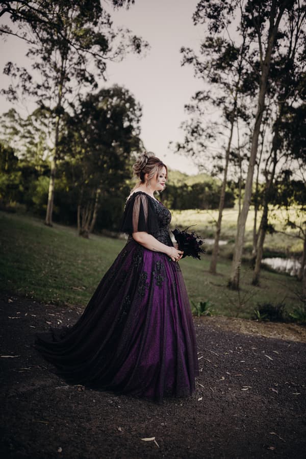 The bride Renae stands alone outdoors at Ocean View Estates wearing a purple and black gown and holding a dark bouquet, with trees and grass in the background.