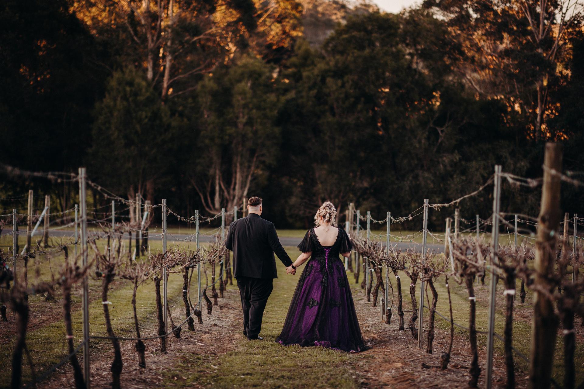 Renae and Adam walk hand in hand down a vineyard row at Ocean View Estates, seen from behind with trees in the background.