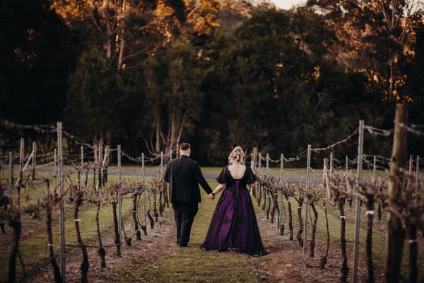Renae and Adam walk hand in hand down a vineyard row, seen from behind with trees in the background. Getting ready photos by She Said Yes.