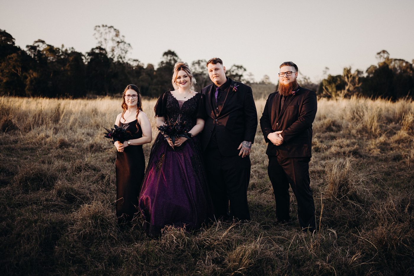 Renae in a dark purple wedding gown and Adam in a black suit stand together with two other individuals dressed in black formal attire in a grassy field. Full day wedding photography by She Said Yes.
