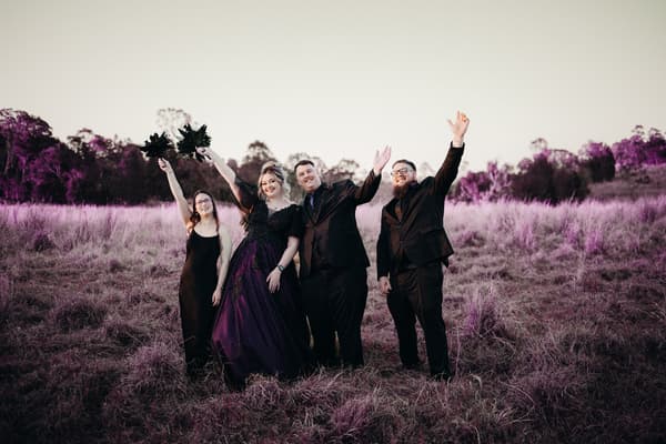 Renae and Adam pose with two attendants in a grassy field, all dressed in formal black attire with the bride in a dark gown, raising their arms in celebration. Wedding photographers Brisbane by She Said Yes.