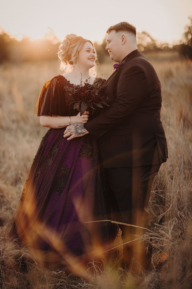 Renae and Adam stand facing each other in a grassy field at Ocean View Estates during their couple portraits session, with Renae holding a dark bouquet and wearing a purple and black dress, and Adam in a black suit.