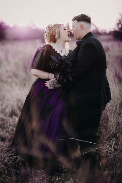 Renae the bride in a purple and black gown and Adam the groom in a black suit stand facing each other in a grassy field, holding a dark bouquet. Sandstone point hotel wedding by She Said Yes.