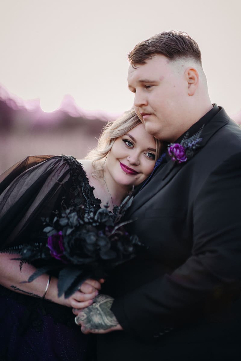 Renae the bride in a black dress with purple lipstick leans her head on Adam the groom's shoulder as they hold hands and a bouquet of dark flowers. Engagment party by She Said Yes.