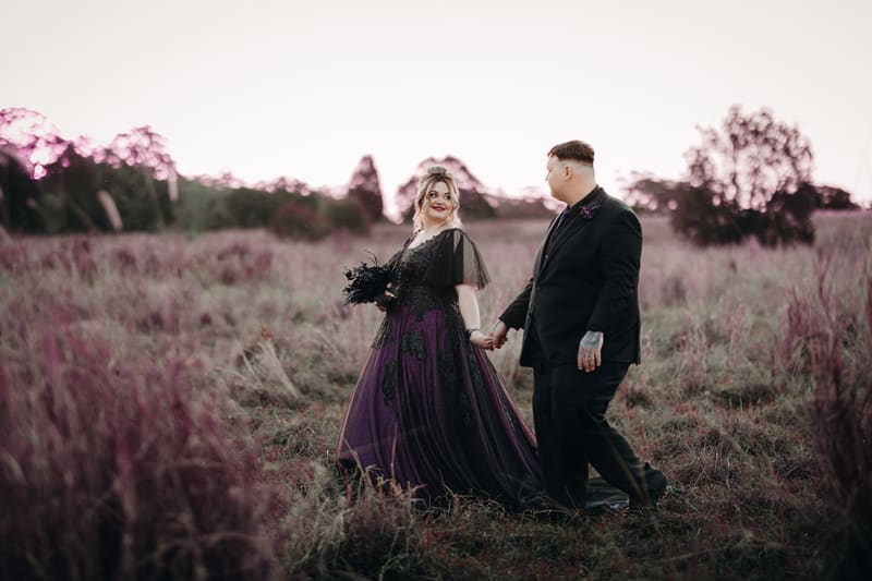 Renae in a dark purple and black wedding gown and Adam in a black suit hold hands while walking through a grassy field at Ocean View Estates during their couple portraits session.