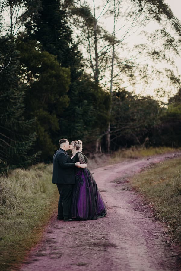 Renae and Adam stand on a dirt path surrounded by trees at Ocean View Estates, sharing a kiss during their couple portraits session.