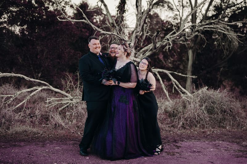 Renae the bride in a purple and black gown and Adam the groom in a black suit pose with two other people in dark attire holding bouquets, standing in front of a large bare tree and dry shrubbery. Wedding photography contracts by She Said Yes.