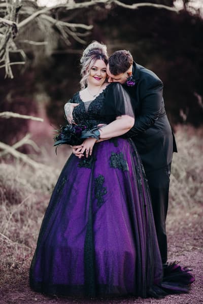 Renae in a purple and black wedding gown holding a bouquet, embraced from behind by Adam in a black suit, posing outdoors. Autumn wedding palette Queensland by She Said Yes.