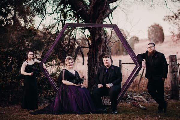 The bride Renae in a dark purple gown and the groom Adam in a black suit sit inside a large wooden hexagonal frame outdoors, accompanied by two attendants dressed in black standing on either side of the frame. Wedding date changed COVID by She Said Yes.