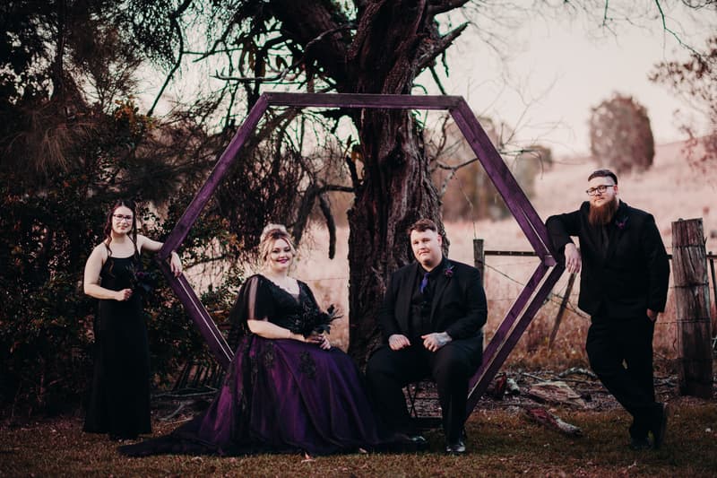 The bride Renae in a dark purple gown and the groom Adam in a black suit sit inside a large wooden hexagonal frame outdoors at Ocean View Estates, accompanied by two attendants dressed in black standing on either side of the frame.
