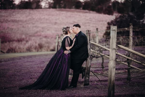 Renae and Adam share a kiss outdoors, with Adam leaning against a wooden fence and Renae wearing a dark purple gown. Event videographer Brisbane by She Said Yes.