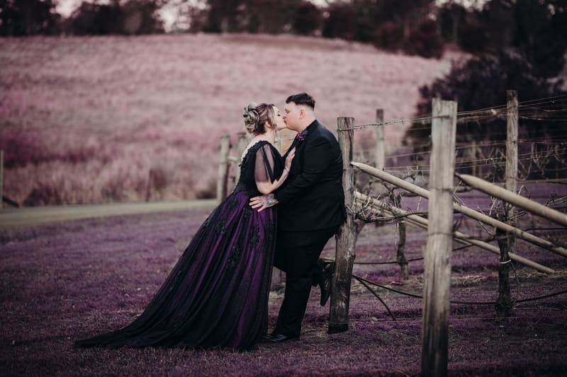 Renae and Adam share a kiss outdoors, with Adam leaning against a wooden fence and Renae wearing a dark purple gown. Brisbane elopement photographer by She Said Yes.