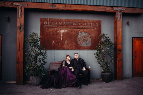 Renae and Adam sit holding hands on a wooden bench beneath a large Ocean View Estates sign flanked by two potted plants.