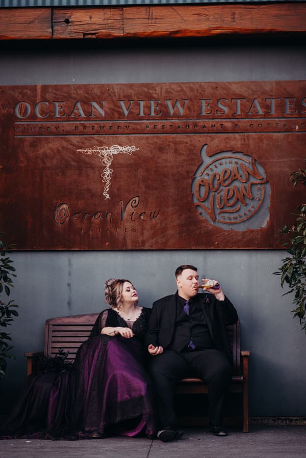 Renae, the bride, in a dark purple gown and Adam, the groom, in a black suit with a purple tie, sit on a wooden bench under a large Ocean View Estates sign, with Adam drinking from a bottle.