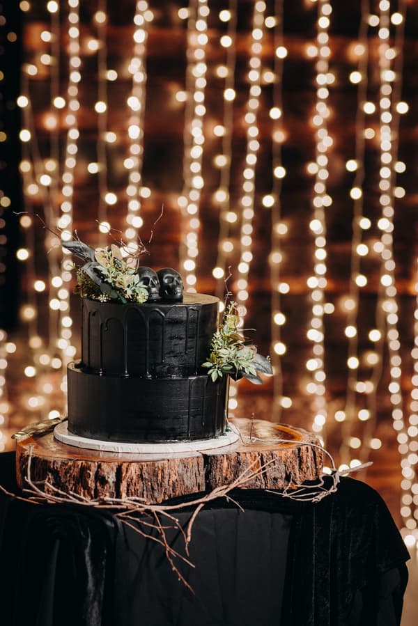 Two-tier black wedding cake decorated with black skull toppers and floral accents, displayed on a wooden slab at Ocean View Estates — Function Room with string lights in the background.