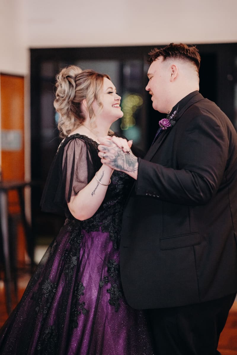 The bride Renae and groom Adam share a dance at the reception in the Ocean View Estates Function Room. Renae wears a purple and black gown with sheer sleeves, and Adam is dressed in a black suit with a purple boutonniere.