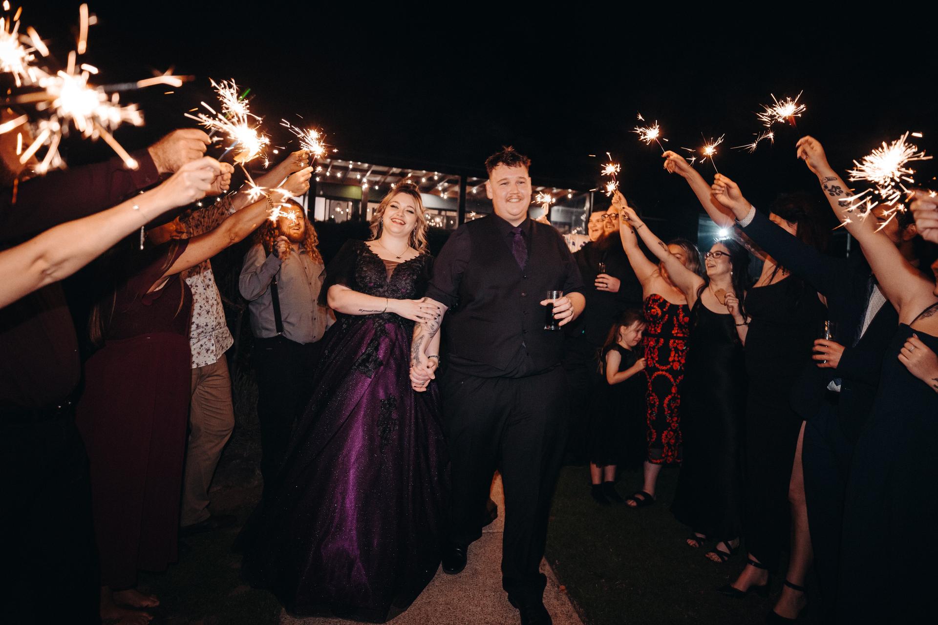 Bride Renae in a dark purple gown and groom Adam in a black shirt and vest walk hand in hand through a pathway lined with guests holding sparklers at Ocean View Estates — Function Room.