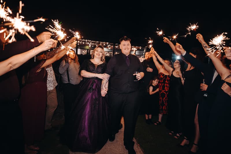 Bride Renae in a dark purple gown and groom Adam in a black shirt and vest walk hand in hand through a pathway lined with guests holding sparklers at Ocean View Estates — Function Room.
