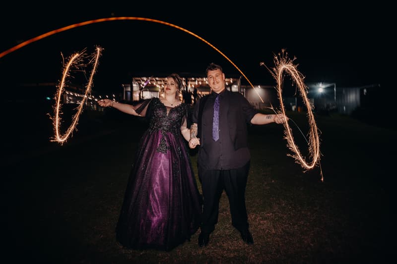 The bride Renae and groom Adam stand outside Ocean View Estates — Function Room at night holding sparklers creating light trails.