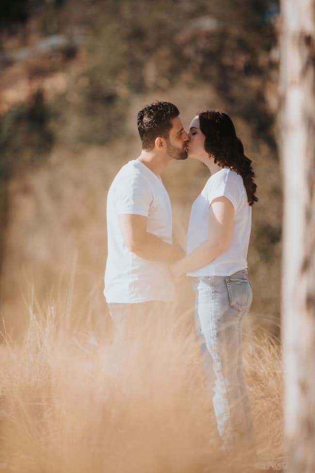 The couple shares a kiss in a sunlit grassy area at Kangaroo Point, Brisbane, wearing casual white tops and jeans.