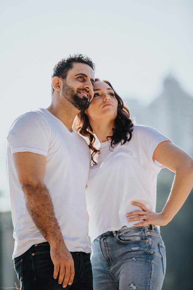The couple stands close, gazing upwards with smiles, dressed in white tops and jeans, against a bright sky.