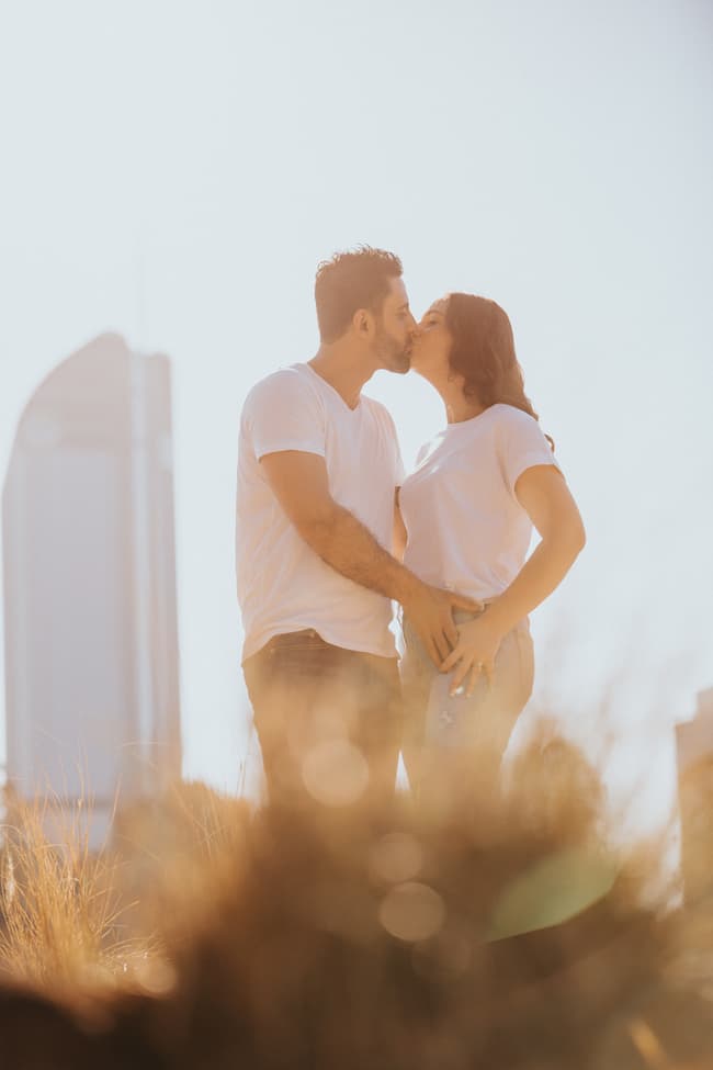 The couple kisses with a tall building in the background, standing in a grassy area at Kangaroo Point.