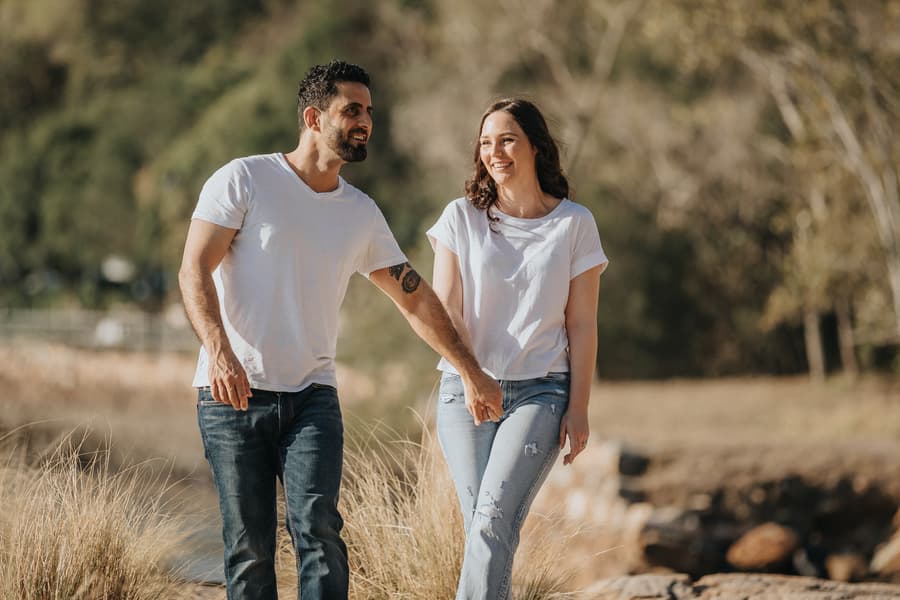The couple smiles and holds hands while walking on a grassy path, surrounded by trees at Kangaroo Point.