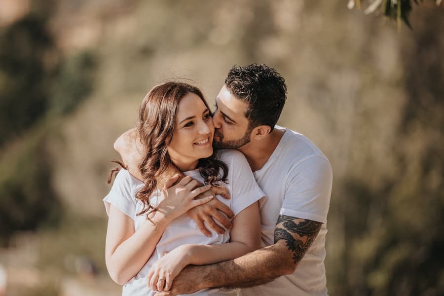 The couple embraces with the man kissing the woman's cheek, both smiling warmly at Kangaroo Point.