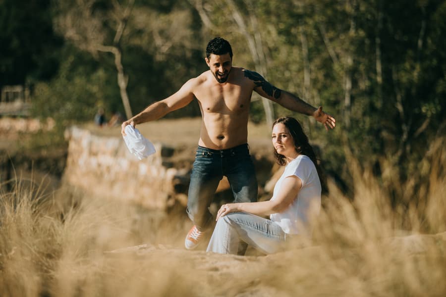The man playfully removes his shirt while the woman sits on a rock, both enjoying a lighthearted moment.