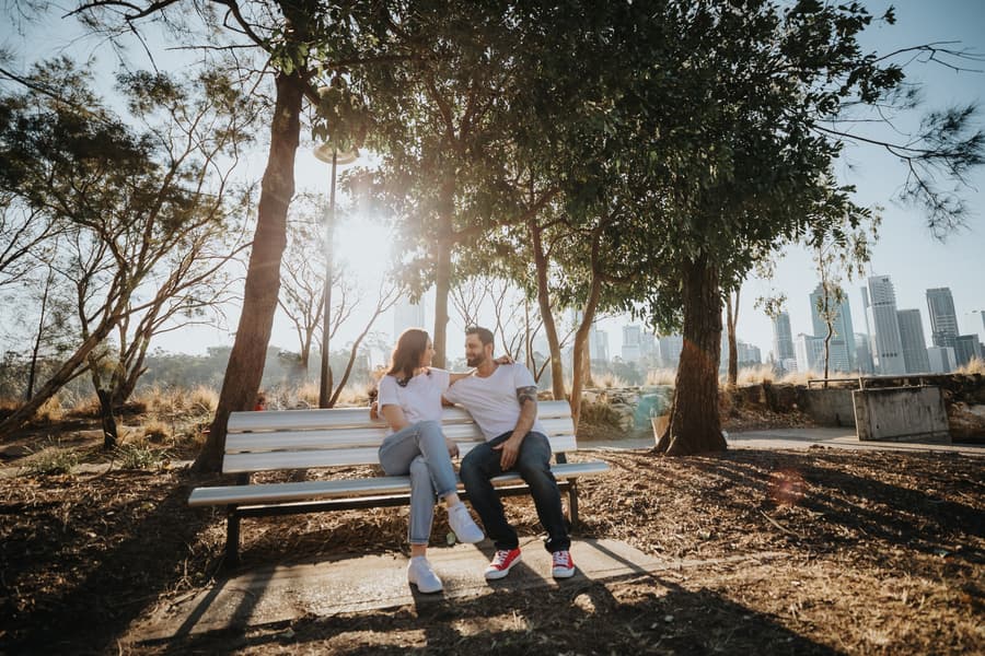 The couple sits on a park bench under a tree, with city buildings in the background at Kangaroo Point.