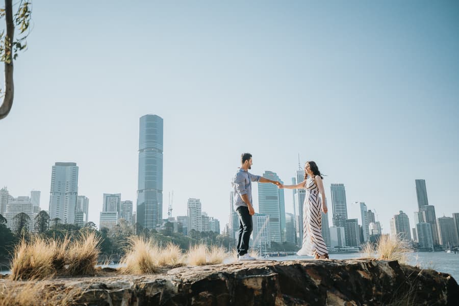The couple stands on a rocky ledge with the Brisbane skyline in the background, holding hands.
