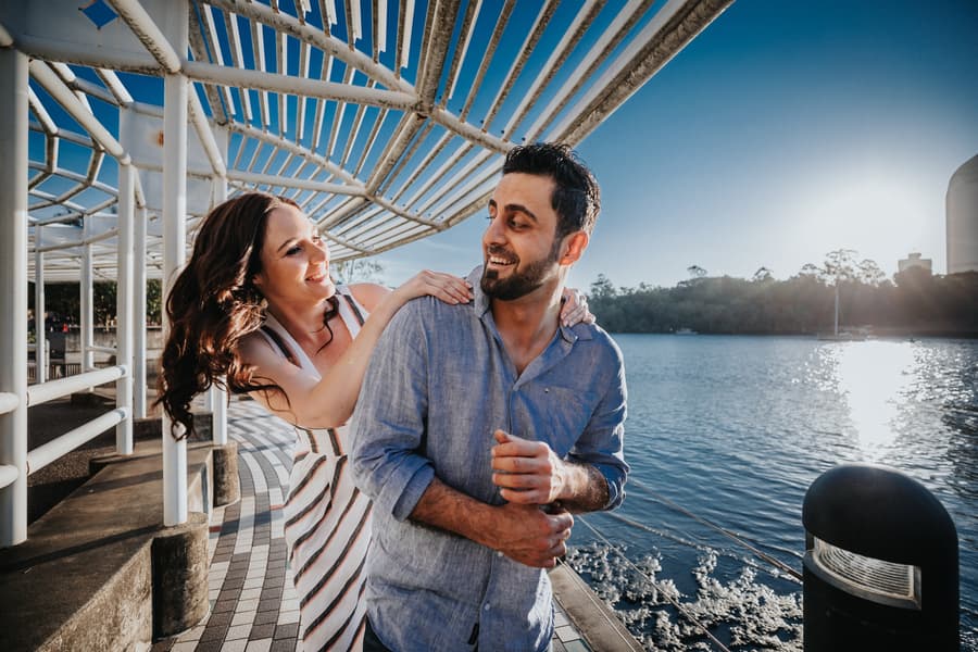 The couple playfully walking along a riverside path at Kangaroo Point, with a bright blue sky above.