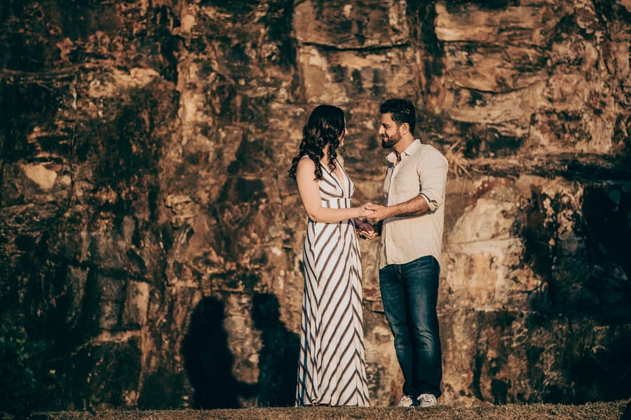 The couple standing close, holding hands in front of a rocky cliff at Kangaroo Point, sharing a tender moment.