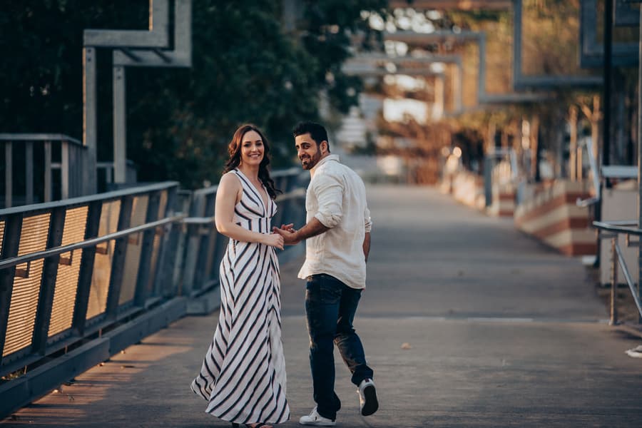 The couple walking hand in hand on a bridge at Kangaroo Point, looking back with smiles.