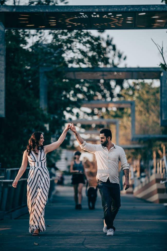The couple holding hands and walking on a bridge at Kangaroo Point, with a person and dog in the background.