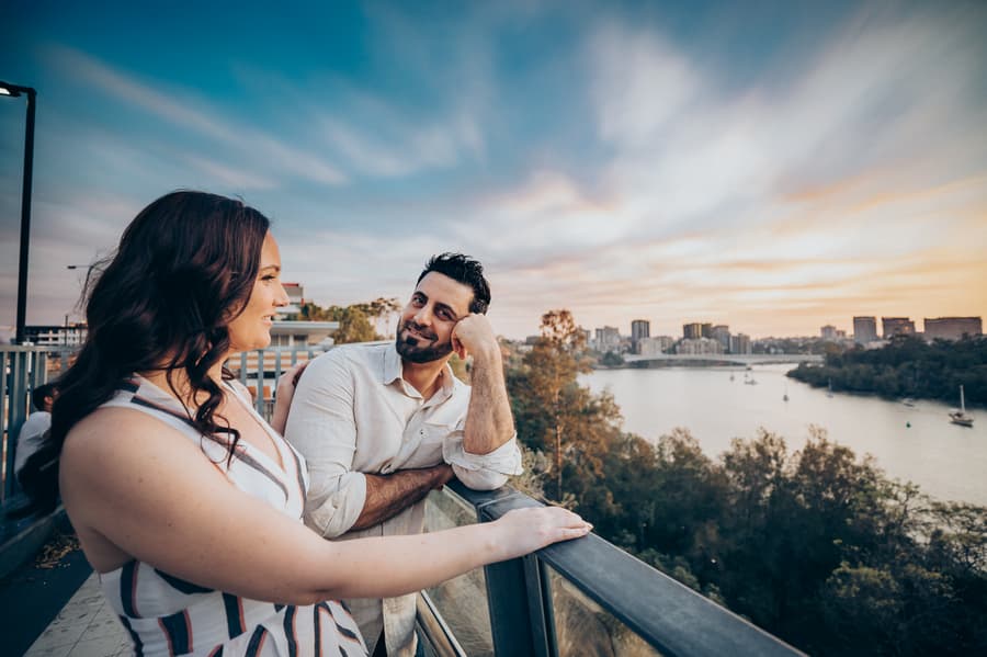 The couple leans on a railing, smiling at each other with a river and cityscape in the background at Kangaroo Point.